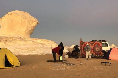 Campement dans le désert blanc égyptien avec des tentes, des personnes et un véhicule devant une formation rocheuse blanche.